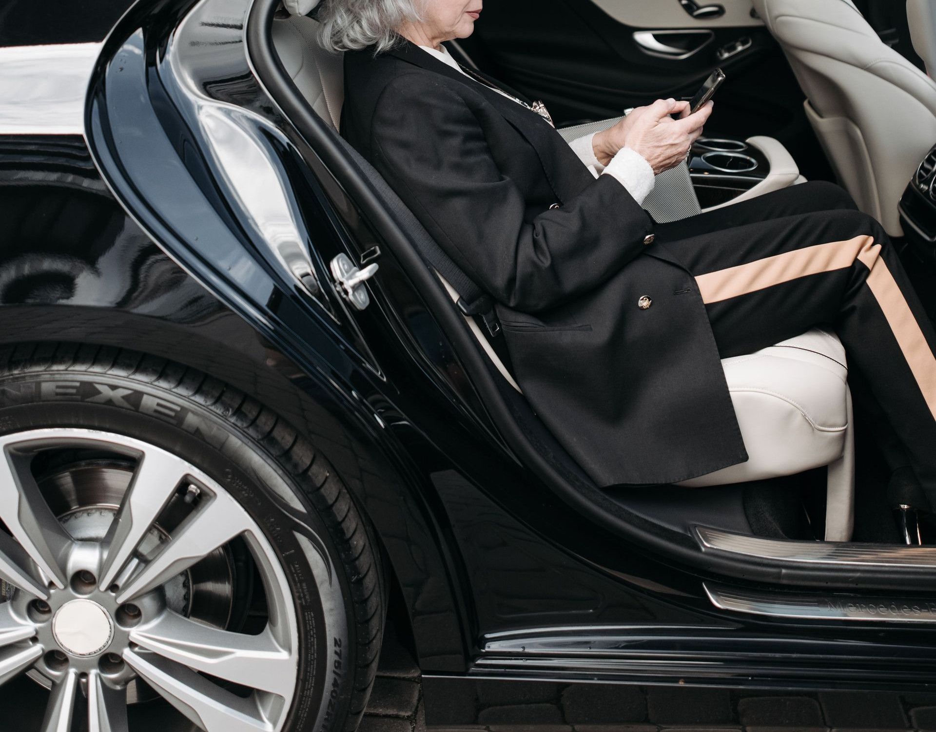 Senior woman in business attire sits in a luxury car, using a tablet outside a modern building.
