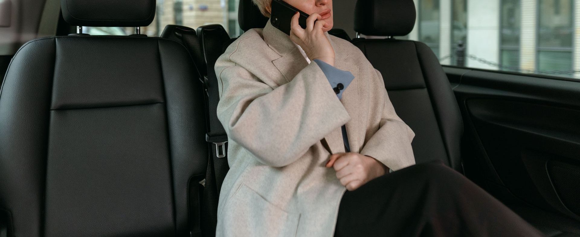 A businesswoman in a coat talks on her smartphone while sitting in the back seat of a car.